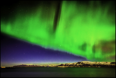 Dayglow-green lights over a dark-blue horizon and tan mountains, all reflected in water — Iceland, February 2014. Photo by Nelson Burack